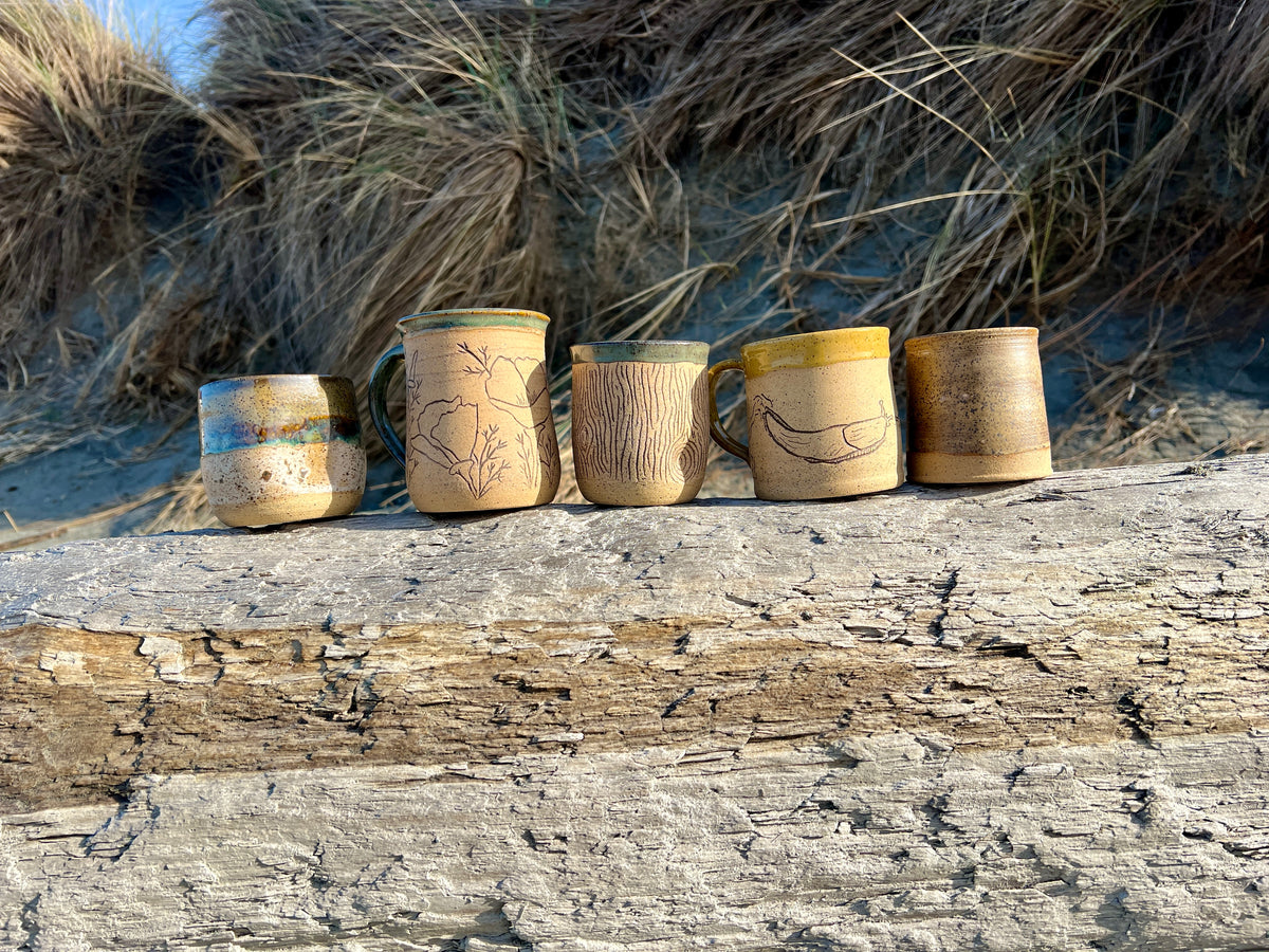 Five handmade ceramic mugs with natural wood-grain patterns and hand-painted designs, lined up on a weathered log with beach grass in the background.